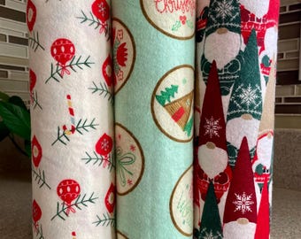 May include: Three rolled Christmas-themed fabric cloths. One cloth is white with red ornaments and green sprigs. Another is light green with circular designs and the text "Merry Christmas". The third cloth has gnomes.