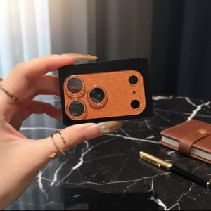 May include: A black and orange phone camera lens protector. The protector has a rectangular shape with three circular lens openings and a glittery orange surface. The item is held by a hand with long, manicured nails.