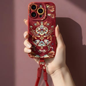 May include: A red phone case with an ornate, traditional design. The case features a detailed pattern in gold, blue, and white, with a matching red lanyard. The phone case has a camera cutout with three lenses.