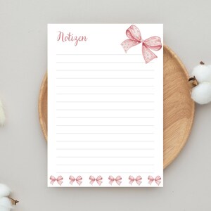 Könnte beinhalten: Ein liniertes Notizblock mit dem Wort "Notizen" in rosa Schrift. Oben rechts befindet sich eine rosa Schleife mit Punkten, und am unteren Rand sind rosa Schleifen angeordnet. Der Notizblock liegt auf einem Holztablett.