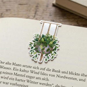 Könnte beinhalten: Dekorativer Lesezeichen mit einer Aquarellillustration einer Hängepflanze. Die Pflanze hat grüne Blätter und befindet sich in einem blau-weiß gemusterten Topf. Das Lesezeichen befindet sich auf einer Buchseite.