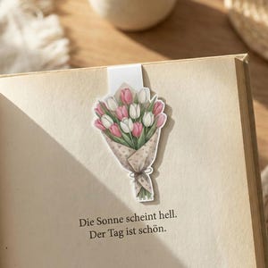 Op de afbeelding: Bloemenbladwijzer met een boeket roze en witte tulpen verpakt in papier met een patroon, geplaatst op een open boek. De Duitse tekst "Die Sonne scheint hell. Der Tag ist schön." is zichtbaar.