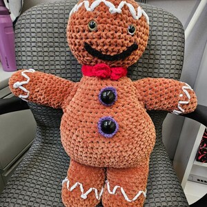 Könnte beinhalten: Handgemachte, plüschige Lebkuchenmann-Puppe mit einem lächelnden Gesicht, schwarzen Augen und einer roten Fliege. Die Puppe hat weiße Zuckergussdetails auf Kopf, Armen und Beinen sowie lila und schwarze Knopfakzente. Sie sitzt auf einem grau gemusterten Stuhl.