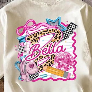 Pode incluir: Uma t-shirt cor de creme com uma impressão gráfica de uma letra B grande com padrão de leopardo e o nome Bella escrito em rosa. O design inclui uma mochila rosa, um lápis amarelo, uma bola de discoteca rosa, estrelas e um laço rosa.