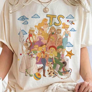K&ouml;nnte beinhalten: Ein cremefarbenes Kurzarm-T-Shirt mit einem gro&szlig;en Grafikdruck mehrerer animierter weiblicher Figuren in verschiedenen Outfits. Das Design enth&auml;lt die Buchstaben TS, eine Rakete, einen gelben Stern-Badge mit der Aufschrift SHERIFF und einen bunten Ball.