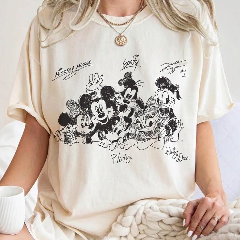 Puede incluir: Una camiseta de color crema con un gr&aacute;fico de l&iacute;neas negras de personajes de Disney, incluyendo a Mickey Mouse, Minnie Mouse, Goofy, Pluto, Donald Duck y Daisy Duck, con sus nombres escritos en cursiva sobre o debajo de cada personaje.