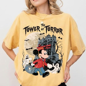 May include: A yellow t-shirt featuring a graphic of Mickey Mouse in a bellhop uniform carrying a suitcase. The design includes the text "TOWER of TORROR," a large moon, a spooky hotel building, bare trees, bats, and two white ghosts.