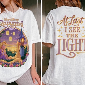 May include: A white t-shirt featuring a front graphic of Rapunzel sitting near a tower with floating lanterns and the text Walt Disney Rapunzel. The back of the shirt displays the text At Last I See The Light in large, stylized purple and gold lettering.