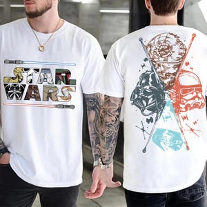 May include: White t-shirt featuring the Star Wars logo with lightsabers and character illustrations. The back of the shirt displays Darth Vader, a Stormtrooper, and the Death Star. The design includes blue, red, and brown accents.