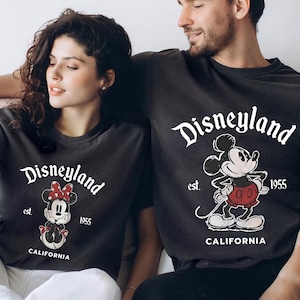 May include: Dark gray t-shirts with the word "Disneyland" in white script. Each shirt features a cartoon character, Mickey or Minnie Mouse, and the text "est. 1955 CALIFORNIA". The shirts are a casual style.