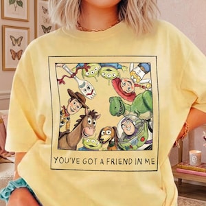 May include: A pale yellow t-shirt featuring a square graphic print of various animated characters including a cowboy, a space ranger, a dinosaur, and a spork. The text "YOU'VE GOT A FRIEND IN ME" is printed in black capital letters below the illustration.
