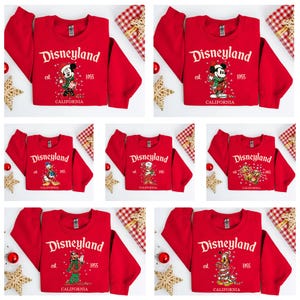 Könnte beinhalten: Eine Kollektion von sieben roten Langarm-Sweatshirts mit dem Schriftzug Disneyland est. 1955 California. Jedes Sweatshirt zeigt eine andere weihnachtliche Disney-Figur, darunter Mickey Maus, Donald Duck und Pluto, umgeben von weißen Schneeflocken.
