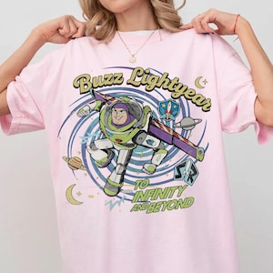 May include: A light pink t-shirt featuring a graphic of Buzz Lightyear in a space-themed swirl. The text reads Buzz Lightyear and To Infinity and Beyond, accompanied by small illustrations of planets, stars, a crescent moon, and a rocket ship emblem.