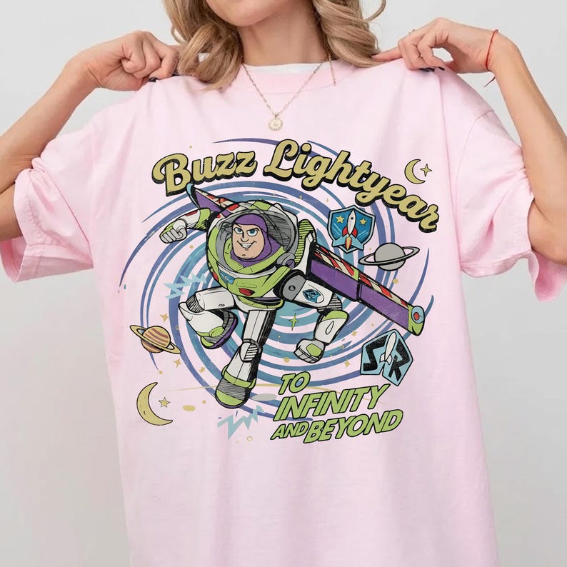 May include: A light pink t-shirt featuring a graphic of Buzz Lightyear in a space-themed swirl. The text reads Buzz Lightyear and To Infinity and Beyond, accompanied by small illustrations of planets, stars, a crescent moon, and a rocket ship emblem.
