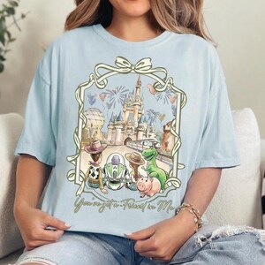 K&ouml;nnte beinhalten: Hellblaues T-Shirt mit einer skurrilen Illustration von Disney-Figuren vor einem Schloss, Feuerwerk und Luftballons. Das Design enth&auml;lt den Text "You've got a Friend in Me". Das Shirt hat eine lockere Passform.
