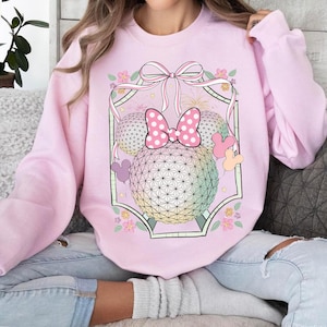 May include: A light pink sweatshirt featuring a graphic of a Minnie Mouse bow and two sphere-shaped heads with a geometric pattern. The bow is pink with white polka dots. The design is framed with a ribbon and floral accents.