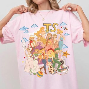 K&ouml;nnte beinhalten: Ein hellrosa T-Shirt mit einem Grafikdruck mehrerer animierter Figuren in verschiedenen Outfits, darunter Weltraum- und Western-Stile. Das Design umfasst blaue Wolken, eine Rakete, einen Sheriff-Stern und die gro&szlig;en gelben Buchstaben TS.