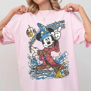 May include: A light pink t-shirt featuring a graphic of Mickey Mouse as a sorcerer wearing a blue star-patterned hat and a red robe. The design includes a yellow broom, buckets of water, and a trail of stars against a splash of blue water.