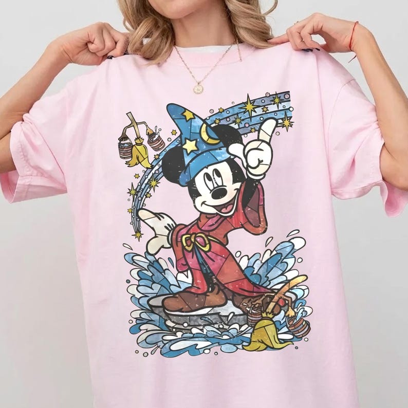 May include: A light pink t-shirt featuring a graphic of Mickey Mouse as a sorcerer wearing a blue star-patterned hat and a red robe. The design includes a yellow broom, buckets of water, and a trail of stars against a splash of blue water.