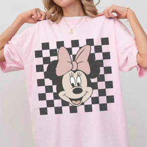May include: A light pink t-shirt featuring a graphic of a cartoon mouse head with a large bow. The design is set against a black and white checkered square background with a distressed, weathered texture throughout the printed artwork.