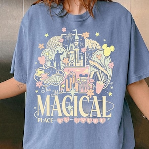 K&ouml;nnte beinhalten: Ein blaues T-Shirt mit einem farbenfrohen Grafikdesign mit einem Schloss, Feuerwerk und anderen Themenparkelementen. Der Text "The Most Magical Place on Earth" ist in einer verspielten Schriftart dargestellt.
