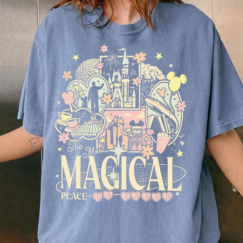 K&ouml;nnte beinhalten: Ein blaues T-Shirt mit einem farbenfrohen Grafikdesign mit einem Schloss, Feuerwerk und anderen Themenparkelementen. Der Text "The Most Magical Place on Earth" ist in einer verspielten Schriftart dargestellt.