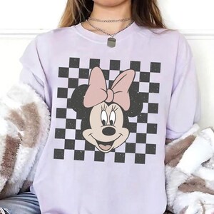 May include: A light purple short-sleeve t-shirt featuring a central graphic of a cartoon mouse head with a large bow, set against a black and white checkered square background. The mouse character has a light pink bow and a cream-colored face.
