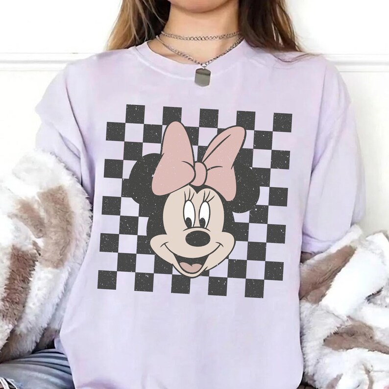 May include: A light purple short-sleeve t-shirt featuring a central graphic of a cartoon mouse head with a large bow, set against a black and white checkered square background. The mouse character has a light pink bow and a cream-colored face.