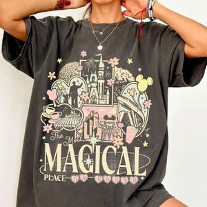 May include: Dark gray t-shirt with a colorful graphic design. The design features a castle, Mickey Mouse, and the text "The Most Magical Place on Earth." The graphic is surrounded by flowers and stars.