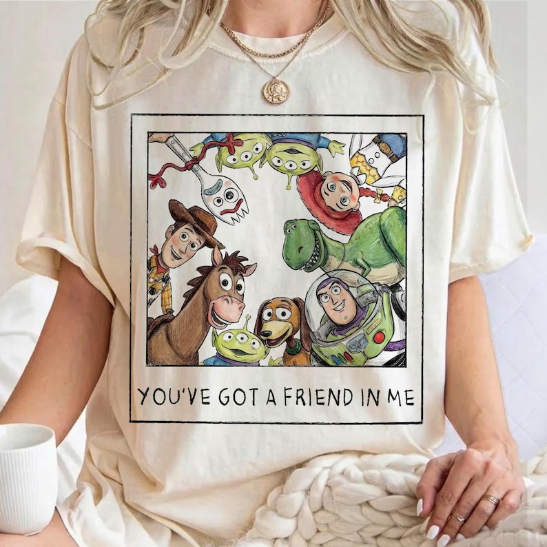 May include: A cream-coloured t-shirt featuring a square graphic print of various animated characters including a cowboy, a space ranger, a dinosaur, and a spork. The text below the characters reads, YOU'VE GOT A FRIEND IN ME.