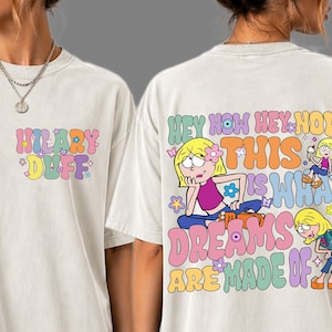 Puede incluir: Una camiseta color hueso con un estampado gráfico. El frente muestra el texto HILARY DUFF en letras de burbuja coloridas. La parte posterior presenta una gran ilustración de un personaje rubio con el texto HEY NOW HEY NOW THIS IS WHAT DREAMS ARE MADE OF.