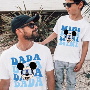 May include: Two white short-sleeved t-shirts featuring a graphic of Mickey Mouse wearing blue-rimmed glasses. One shirt displays the word DADA in a repeating blue wavy font, while the other shirt displays the word MINI in a matching blue wavy font.
