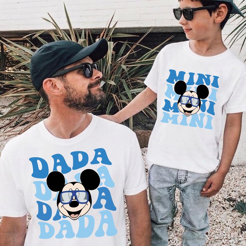 May include: Two white short-sleeved t-shirts featuring a graphic of Mickey Mouse wearing blue-rimmed glasses. One shirt displays the word DADA in a repeating blue wavy font, while the other shirt displays the word MINI in a matching blue wavy font.