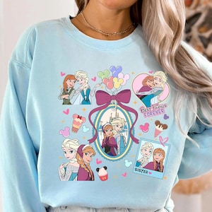 Könnte beinhalten: Hellblaues Sweatshirt mit Illustrationen von zwei animierten weiblichen Figuren. Das Design enthält den Text "Best Friends Forever" und "Sister", zusammen mit Herzen, Eiswaffeln und Luftballons. Das Sweatshirt hat einen Rundhalsausschnitt und lange Ärmel.