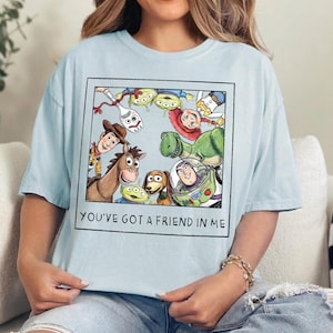 May include: A light blue t-shirt featuring a square graphic print of various animated characters including a cowboy, a space ranger, a dinosaur, and a spork. The text "YOU'VE GOT A FRIEND IN ME" is printed in black capital letters below the illustration.