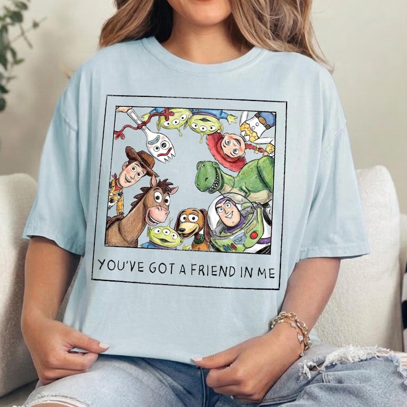 May include: A light blue t-shirt featuring a square graphic print of various animated characters including a cowboy, a space ranger, a dinosaur, and a spork. The text "YOU'VE GOT A FRIEND IN ME" is printed in black capital letters below the illustration.