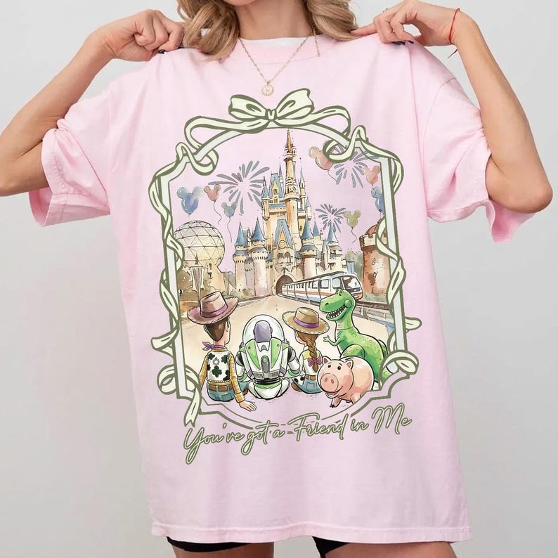 K&ouml;nnte beinhalten: Hellrosa T-Shirt mit einem farbenfrohen Grafikdesign von Toy Story-Charakteren vor einem Schloss. Das Design enth&auml;lt den Text "You've got a Friend in Me" in gr&uuml;ner Schrift, umrahmt von einem dekorativen Rahmen.
