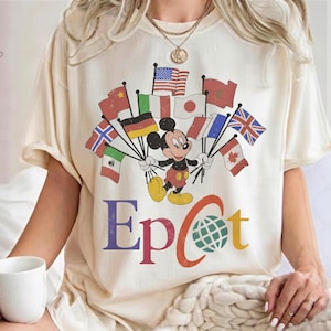 May include: A cream-colored t-shirt featuring a graphic of Mickey Mouse holding a cluster of various international flags. Below the character, the text reads Epcot in colorful, bold lettering, with the letter O replaced by a green globe icon.