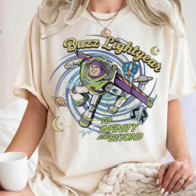 May include: A cream-coloured t-shirt featuring a graphic of Buzz Lightyear in a space-themed design with blue swirls, planets, and stars. The text reads Buzz Lightyear TO INFINITY AND BEYOND in green and black lettering with a small rocket ship emblem.