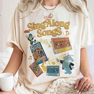May include: Cream-colored t-shirt featuring the text "Disney Sing Along Songs" in yellow and orange. The shirt includes illustrations of Disney characters, cassette tapes, and musical notes, evoking a vintage aesthetic.