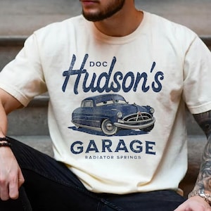 Peut inclure: T-shirt crème avec une voiture de dessin animé bleue et le texte "Doc Hudson's Garage, Radiator Springs." Le design comprend le nom de la voiture au-dessus de l'image et le nom du garage en dessous. Le t-shirt a des manches courtes.