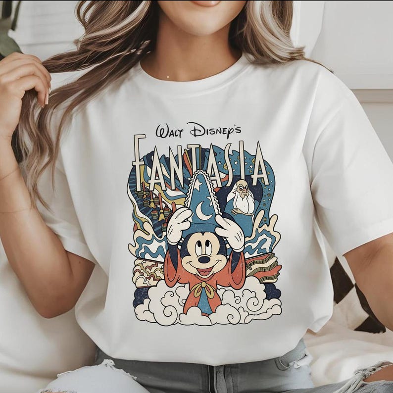 以下が含まれることがあります： ウォルト・ディズニーのファンタジアのカラフルなグラフィックが特徴の白いTシャツ。デザインには、魔法使いの帽子をかぶったミッキーマウスが、青、赤、黄色の様式化されたイメージに囲まれて描かれています。「Walt Disney's FANTASIA」の文字がグラフィックの上に表示されています。
