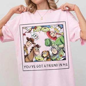 May include: A light pink t-shirt featuring a square graphic print of various animated characters including a cowboy, a space ranger, a dinosaur, a dog, and aliens. The text "YOU'VE GOT A FRIEND IN ME" is printed in black at the bottom of the frame.