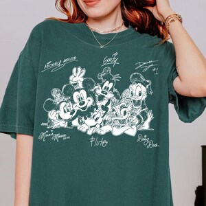 Puede incluir: Una camiseta de manga corta de color verde oscuro con un gr&aacute;fico de l&iacute;neas blancas de Mickey Mouse, Minnie Mouse, Goofy, Pluto, Donald Duck y Daisy Duck. Los nombres de los personajes est&aacute;n escritos en letra cursiva alrededor.