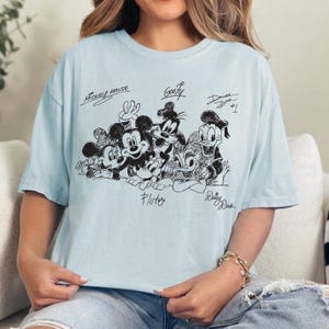Puede incluir: Una camiseta azul claro con un gr&aacute;fico de l&iacute;neas negras de personajes de Disney, incluyendo a Mickey Mouse, Minnie Mouse, Goofy, Pluto, Donald Duck y Daisy Duck. Los nombres de los personajes est&aacute;n escritos en cursiva sobre o bajo ellos.