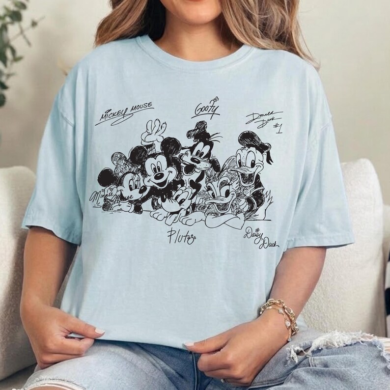 Puede incluir: Una camiseta azul claro con un gr&aacute;fico de l&iacute;neas negras de personajes de Disney, incluyendo a Mickey Mouse, Minnie Mouse, Goofy, Pluto, Donald Duck y Daisy Duck. Los nombres de los personajes est&aacute;n escritos en cursiva sobre o bajo ellos.