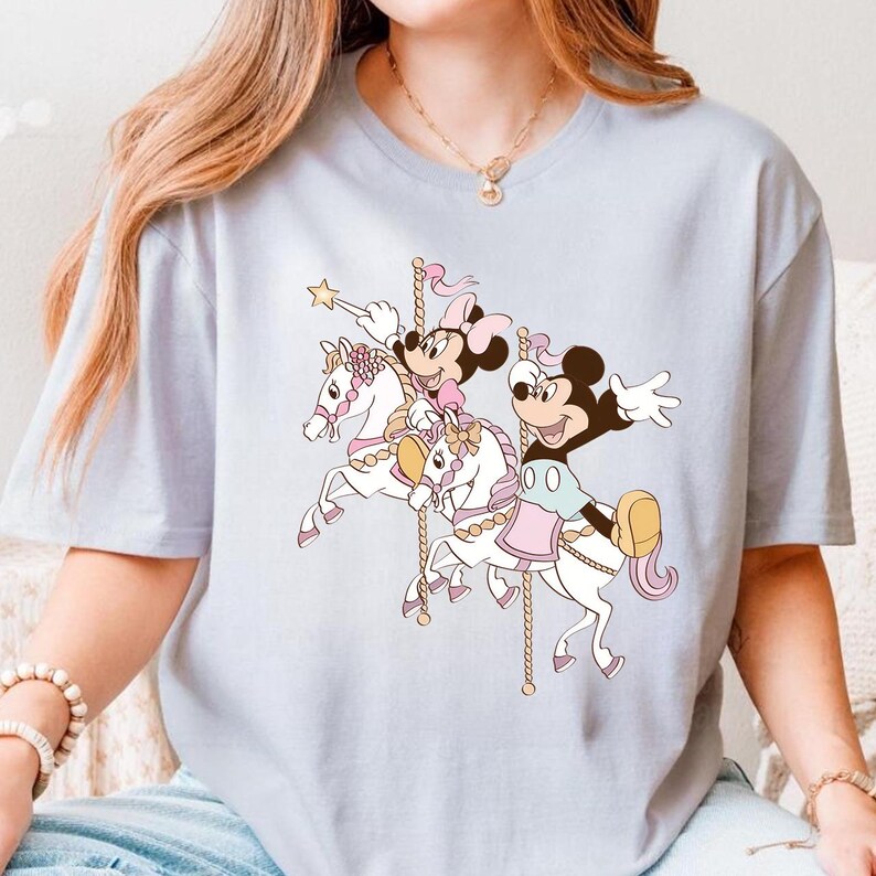 Pode incluir: Camiseta azul clara com um desenho de carrossel com Mickey e Minnie Mouse. Mickey usa um colete azul e Minnie um la&ccedil;o rosa. Os cavalos do carrossel s&atilde;o brancos com detalhes rosa e dourado.