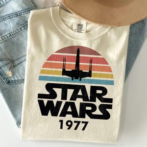 Könnte beinhalten: Ein cremefarbenes Kurzarm-T-Shirt mit einer Grafik der schwarzen Silhouette eines X-Wing-Sternenjägers vor einem Retro-Sonnenuntergang mit roten, orangen, gelben und blauen Streifen. Der Text STAR WARS und 1977 ist darunter schwarz gedruckt.