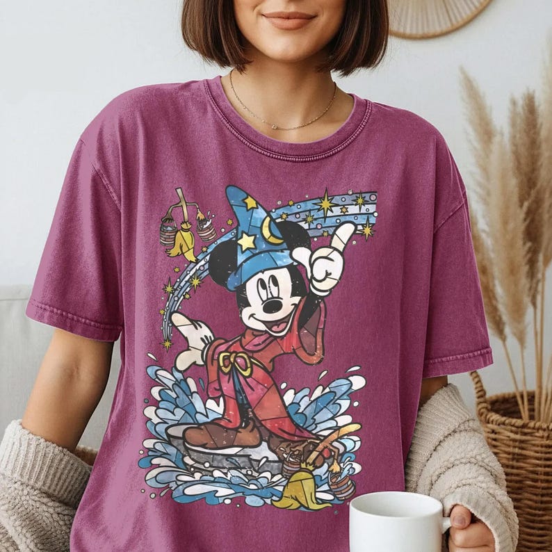 May include: A dark magenta short-sleeve t-shirt featuring a graphic print of Mickey Mouse as a sorcerer wearing a blue star-patterned hat and red robe, standing amidst blue water splashes with yellow brooms and a trail of stars in the background.