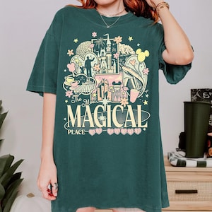K&ouml;nnte beinhalten: Dunkelgr&uuml;nes T-Shirt mit einem farbenfrohen Aufdruck mit dem Text "The Most Magical Place on Earth". Die Grafik zeigt ein Schloss, Micky Maus und andere Themenparkelemente. Das Shirt hat kurze &Auml;rmel und einen Rundhalsausschnitt.